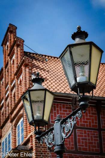 Alte Laternen vor Backsteinfassade  Lüneburg Niedersachsen Deutschland by Peter Ehlert in Dieses detailreiche Foto zeigt zwei kunstvoll verzierte, historische Straßenlaternen im Vordergrund, die vor einer traditionellen Backsteinfassade mit Treppengiebel und roten Dachziegeln in Lüneburg stehen. Die klare Komposition und das helle Tageslicht